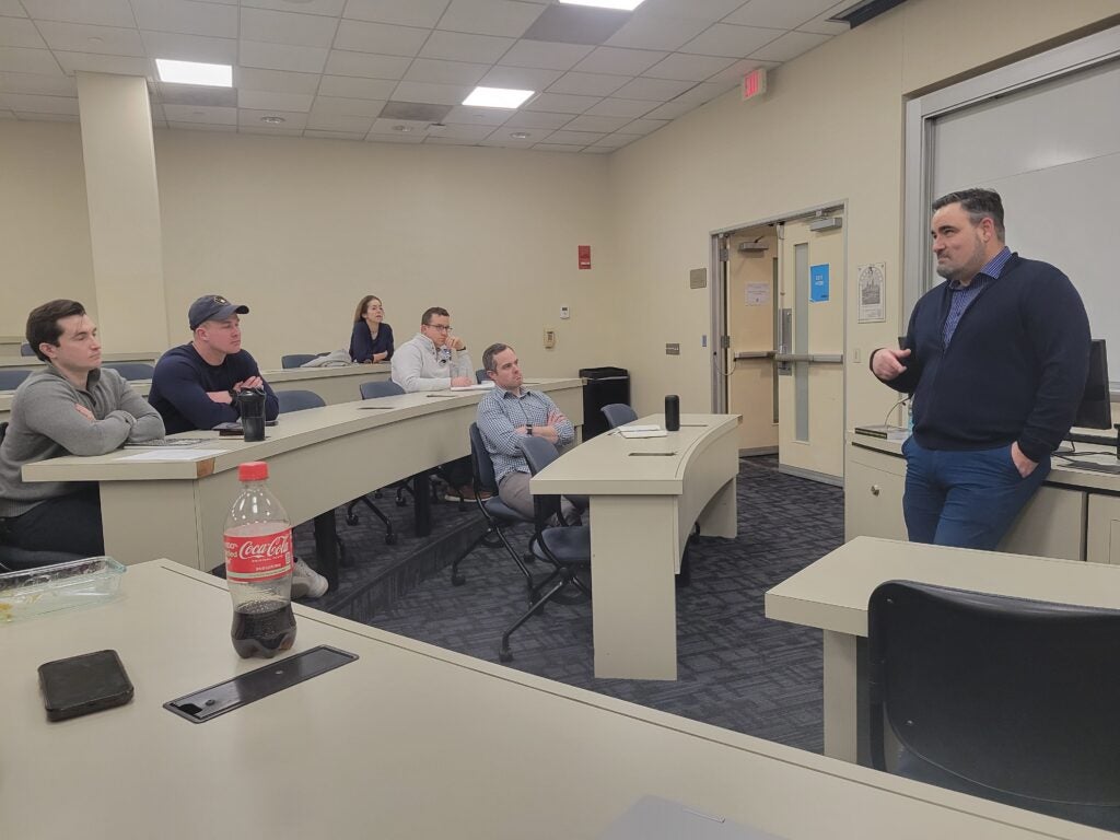 A man stands at the front of a classroom, speaking to six attentive students seated in rows. The room is brightly lit and has a professional atmosphere.