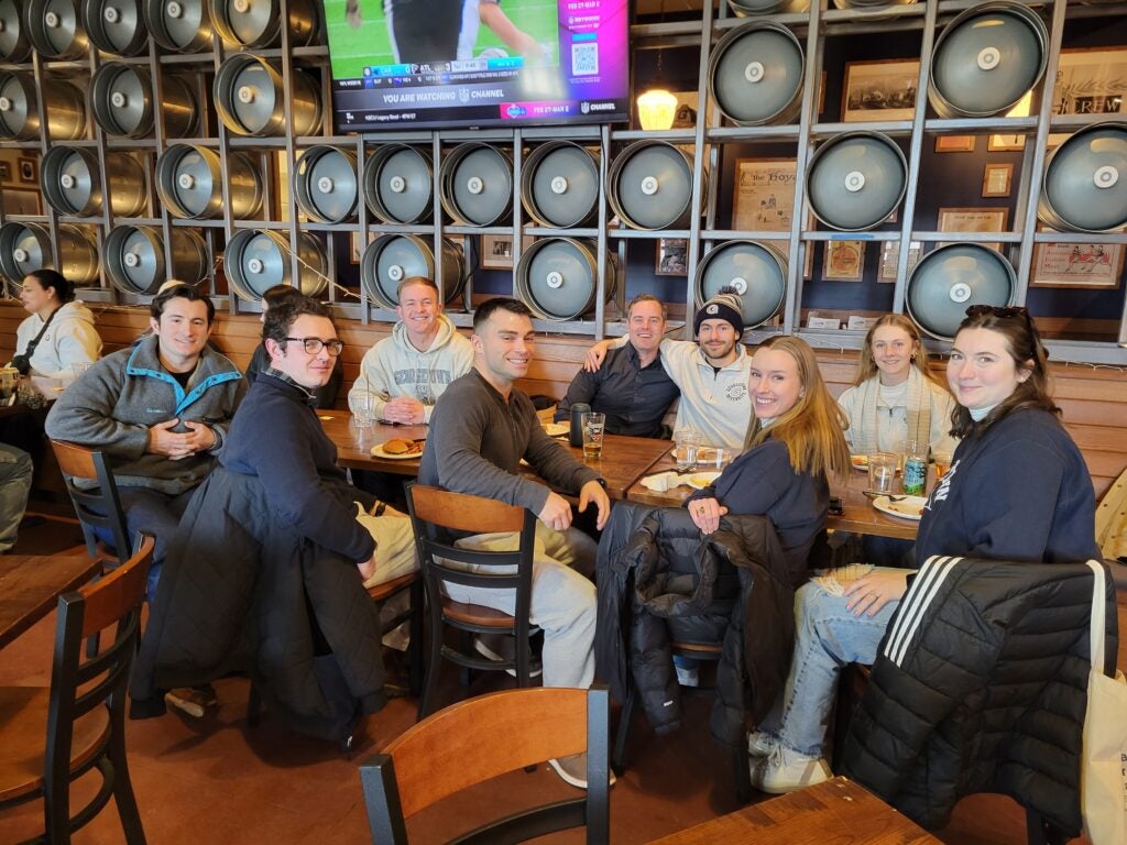 A group of nine people smiles, seated at a wooden table in a lively restaurant with barrel decorations. A TV above shows a football game. 