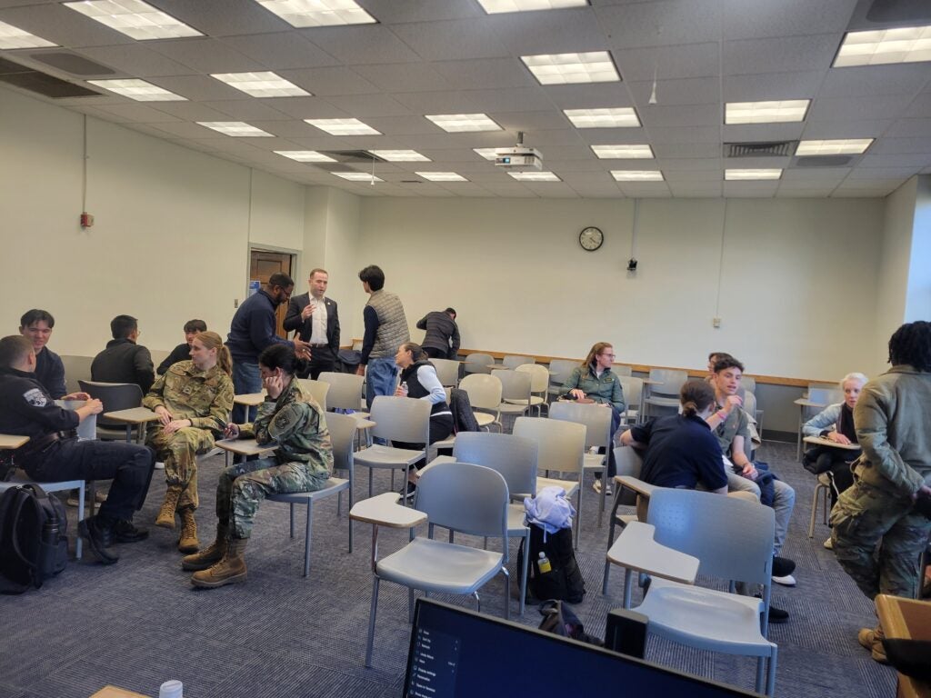 A classroom scene with a diverse group of people in conversation, some in military uniform. Desks in rows fill the room.