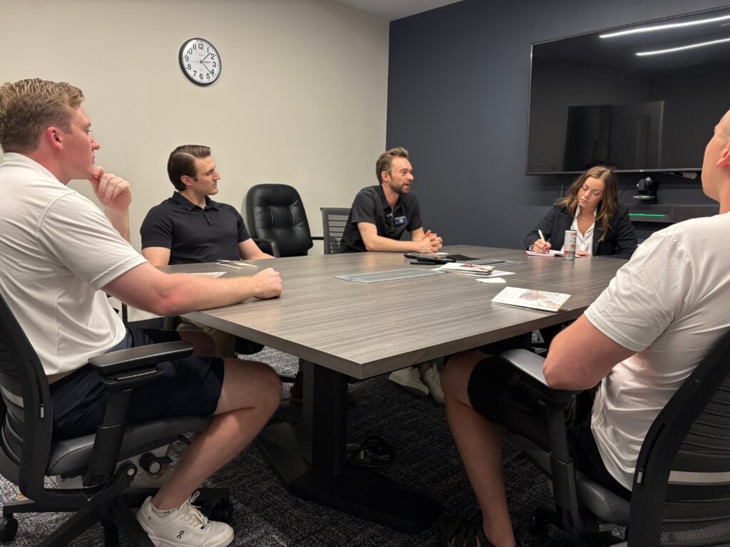 A group of five people sit around a conference table in a meeting room. A woman is taking notes while others appear engaged.