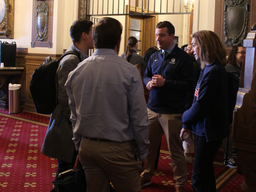 Four individuals are engaged in conversation in an ornate room with red carpet and wooden decor.