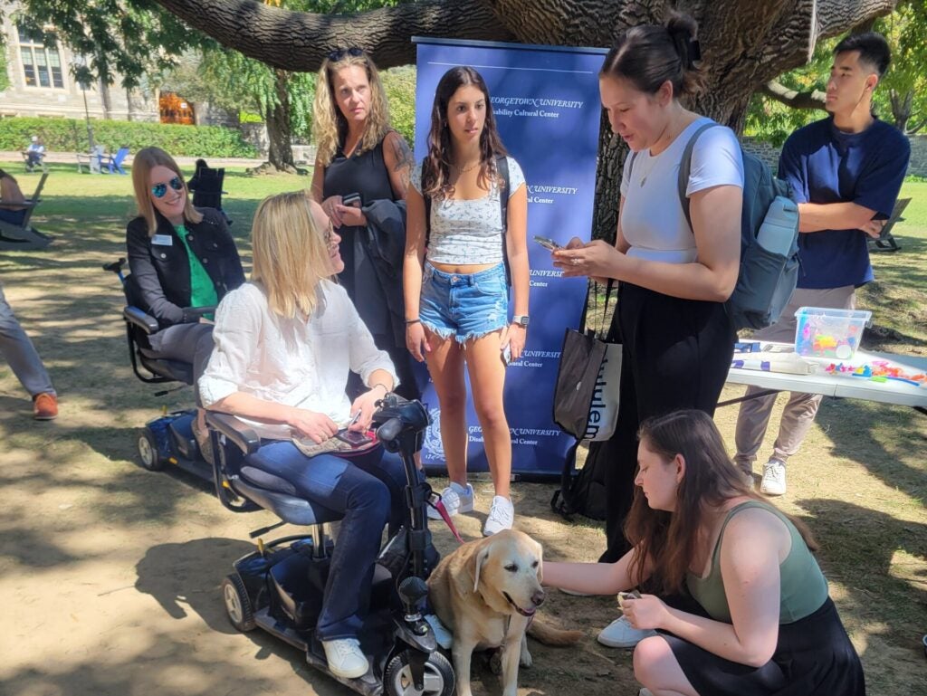 A group of people gathers outdoors around a woman in a wheelchair with a golden retriever. They appear engaged and happy. A blue banner is in the background.