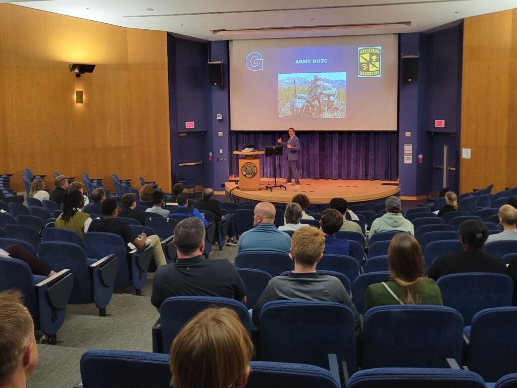 Audience members sit in a lecture hall, listening to a speaker on stage. A large screen displays "Army ROTC" with related imagery
