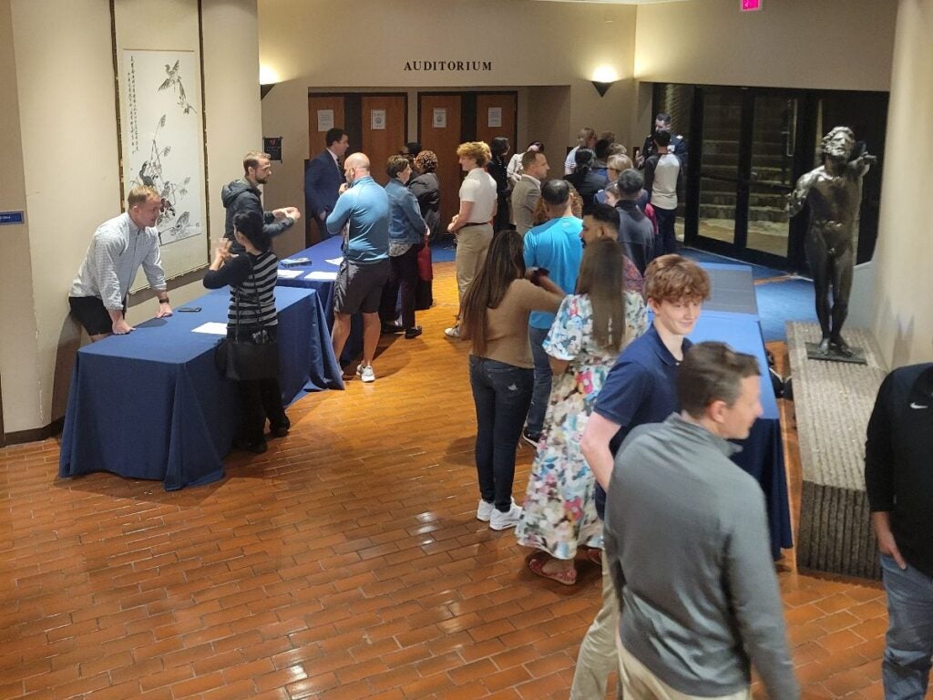 A group of people stands in line outside an auditorium. Tables with blue cloths are on the left, and a metallic statue is on the right.