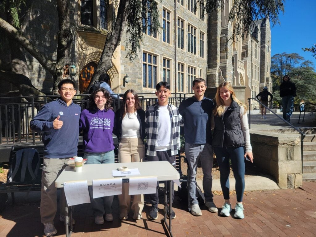 A group of six people smiling outdoors, standing behind a table with a sign labeled "GUAFA" and a historic stone building in the background.