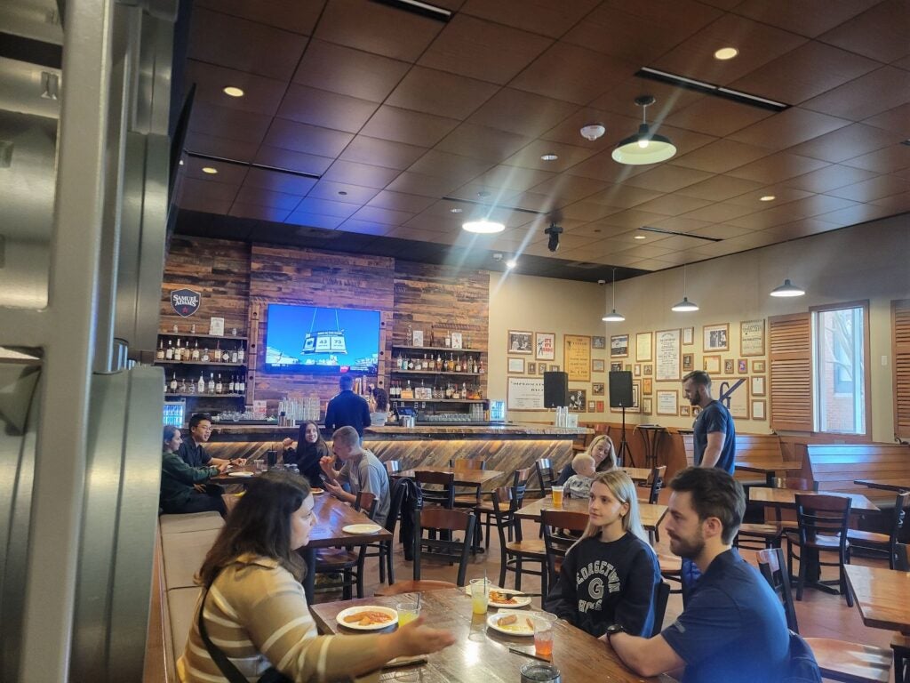 Casual dining scene in a rustic bar with wooden decor. Patrons enjoy meals and conversations. A bartender is at work with a TV in the background.