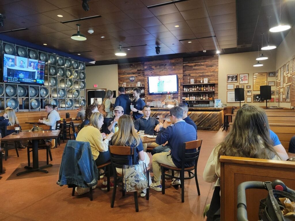 A lively brewery scene shows groups of people socializing at wooden tables. The rustic decor includes wall-mounted barrels and a central TV. 