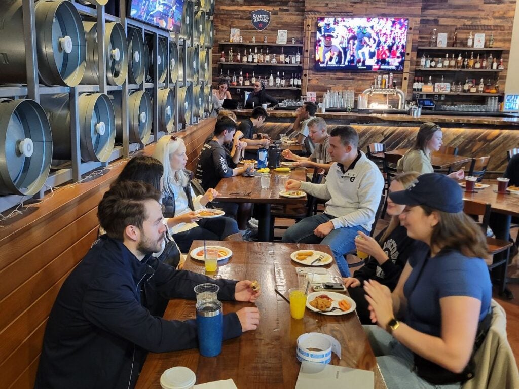 A lively bar scene with people seated at a long table, enjoying food and drinks. Two large TVs display sports games.