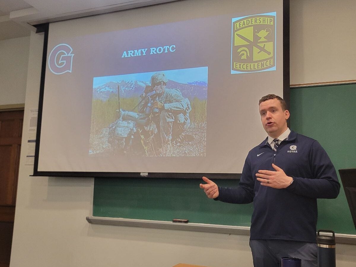 A man presents a slideshow on Army ROTC in a classroom, gesturing towards the screen displaying military imagery and leadership symbols.