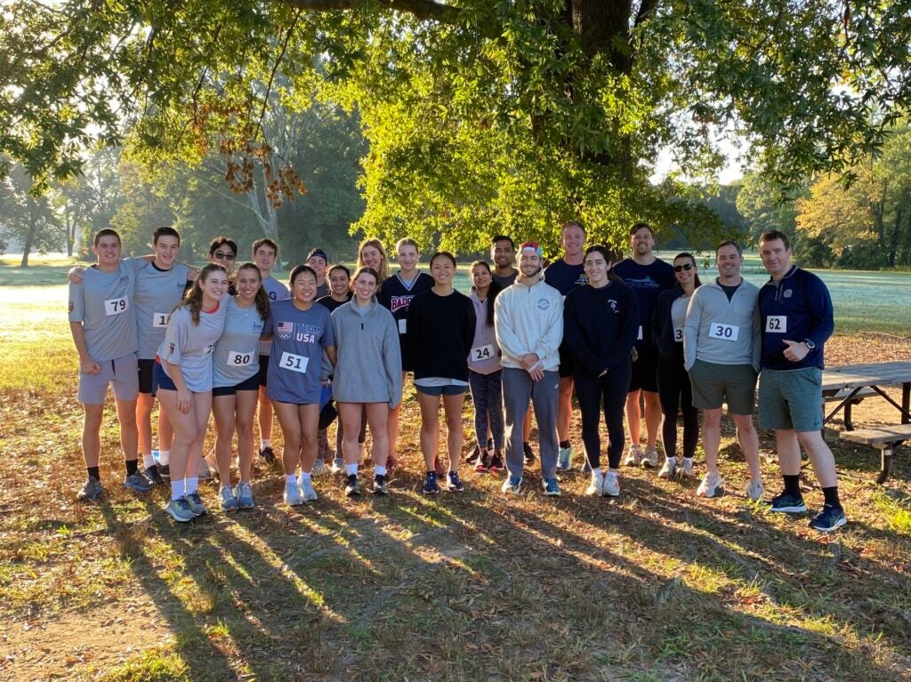 A group of smiling runners stands together under a large tree in a sunlit park. They're wearing race bibs, conveying a sense of camaraderie and achievement.
