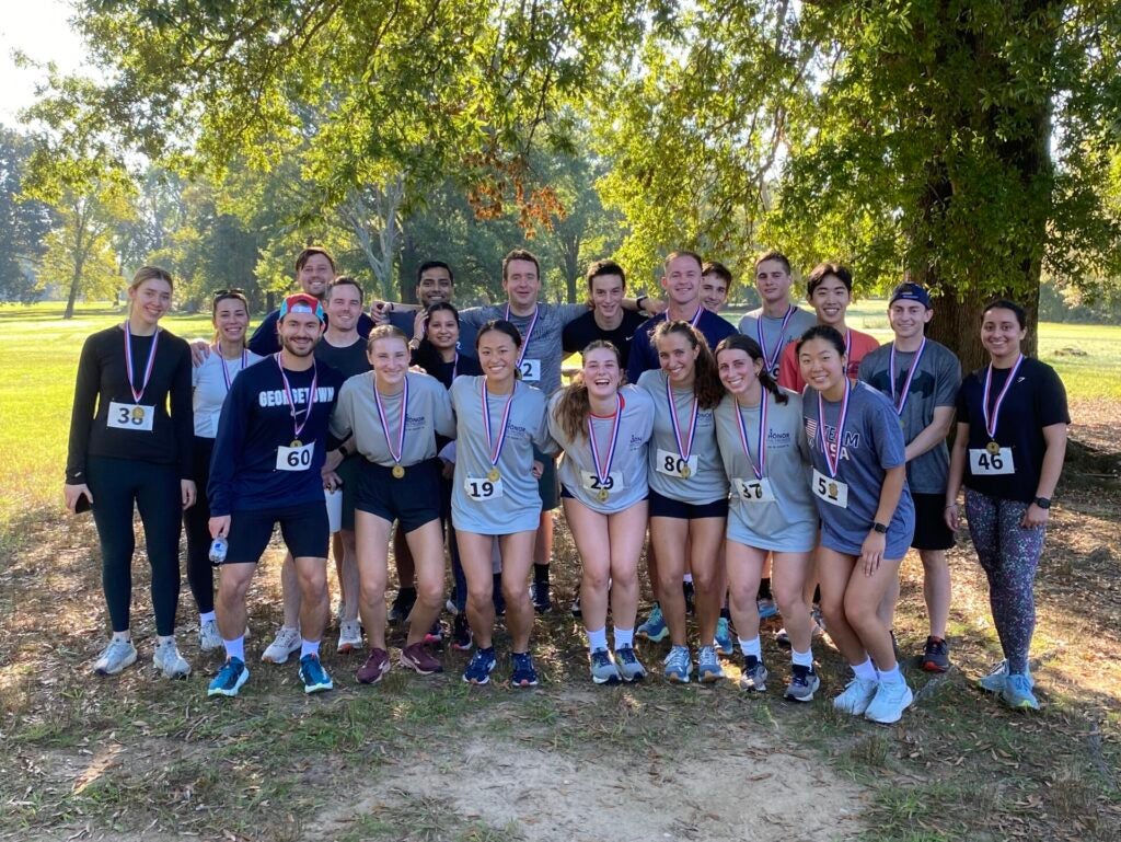 A group of smiling runners wearing medals and numbered race bibs pose under a large tree in a sunny park, exuding joy and camaraderie.
