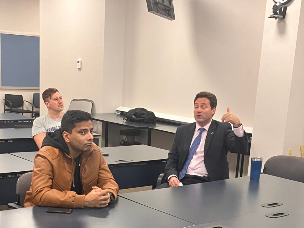 A man in a suit is speaking animatedly to two others in a conference room. One man listens attentively, while the other looks thoughtful.