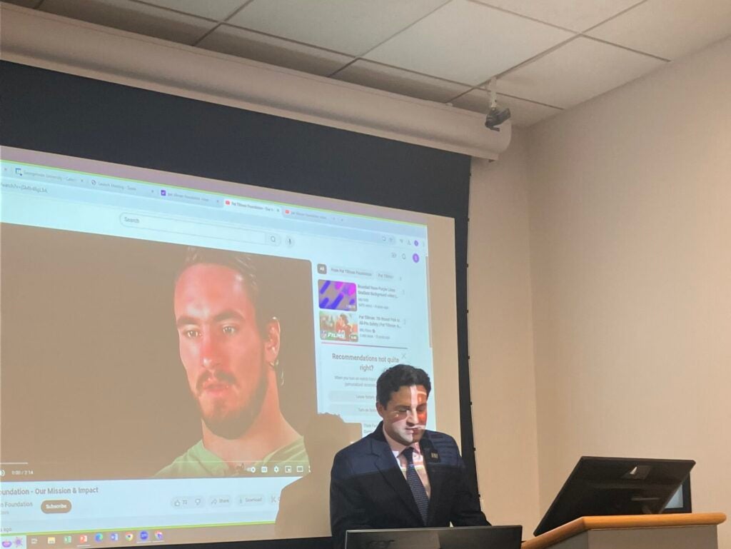 Man in a suit presents in a classroom, standing at a podium with a computer. A video of another man is projected on the screen behind him.