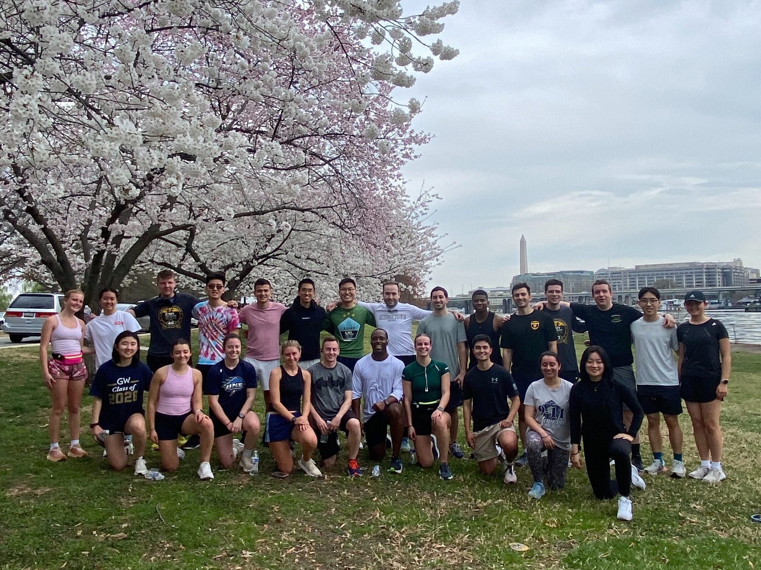 A group of 26 people poses cheerfully under blooming cherry blossom trees. The scene is bright, with a cloudy sky and a distant monument visible.