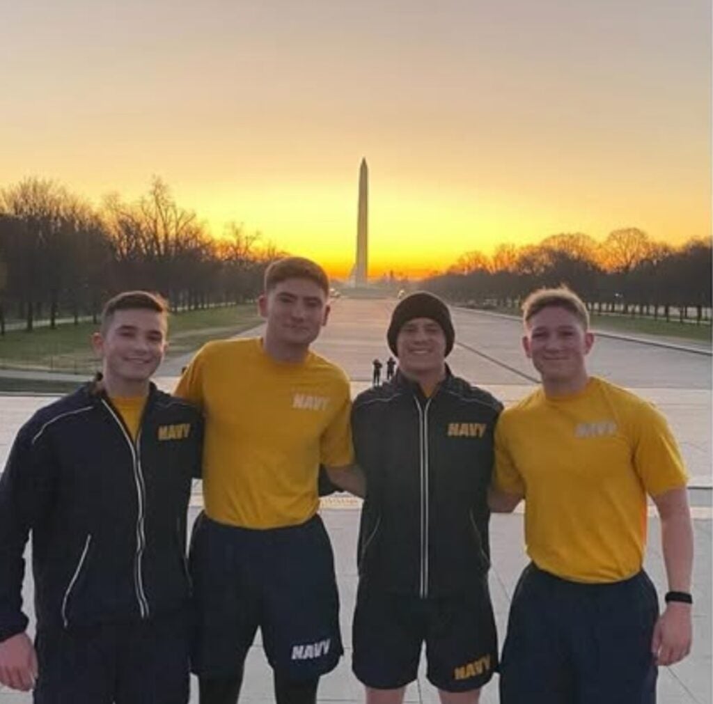 Four people in Navy gear stand arm-in-arm, smiling, with the Washington Monument in the background against a vibrant sunrise.