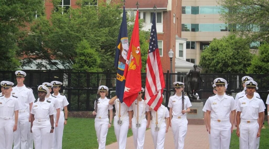 A group of uniformed naval officers stand in formation outdoors, holding flags including the American flag. The setting is a park with greenery and a statue.