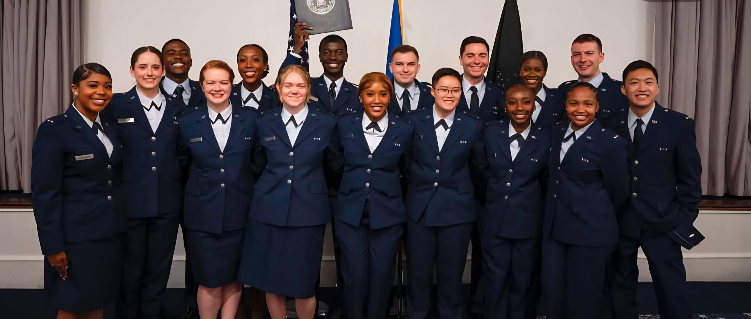 A diverse group of 15 uniformed military personnel smile as they pose in two rows. One person holds a certificate, conveying pride and unity.