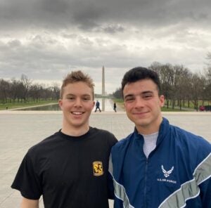 Two young men smile warmly in front of the Washington Monument on a cloudy day, with a reflection pool and bare trees in the background.