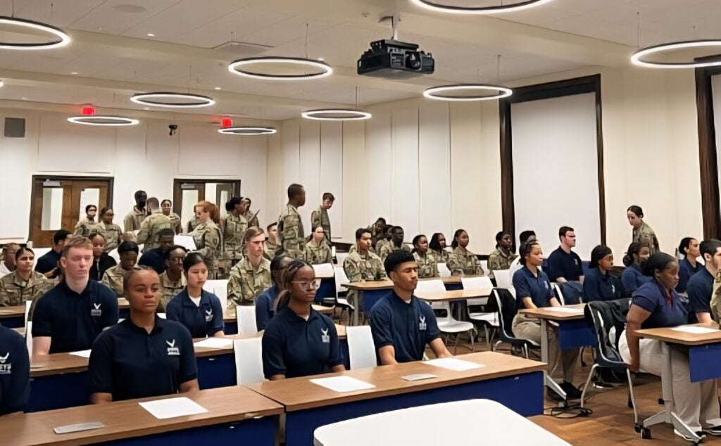 A classroom full of students in Air Force uniforms sit at desks and others in military attire stand. Modern circular ceiling lights illuminate the room.