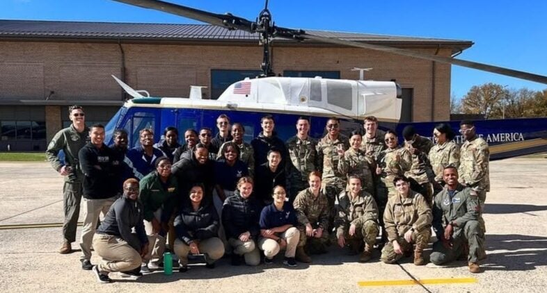 Group of diverse individuals in military and casual attire pose smiling in front of a blue helicopter, on a sunny day with a building in the background.