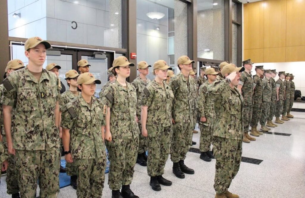 A group of uniformed military personnel in camouflage stand in formation indoors. One salutes, conveying a sense of order and discipline.