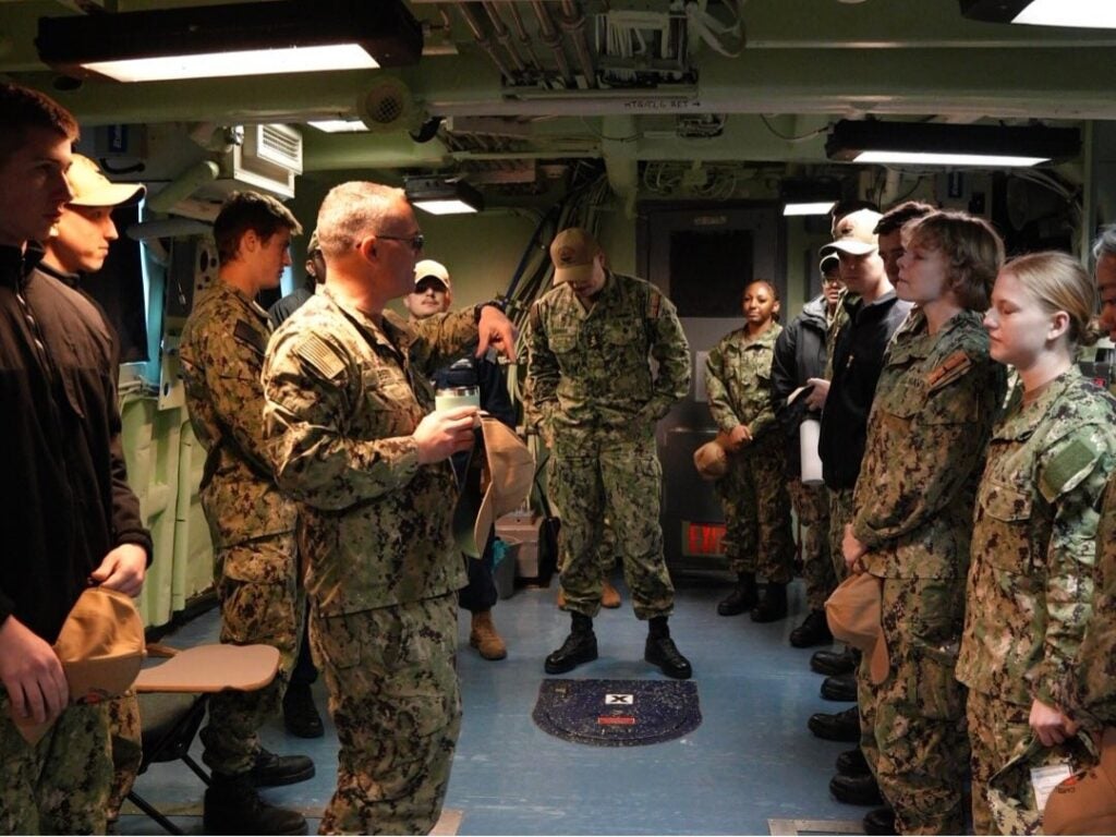 Military personnel in camouflage uniforms stand attentively in a dimly lit room, receiving instructions from a senior officer holding a cap.