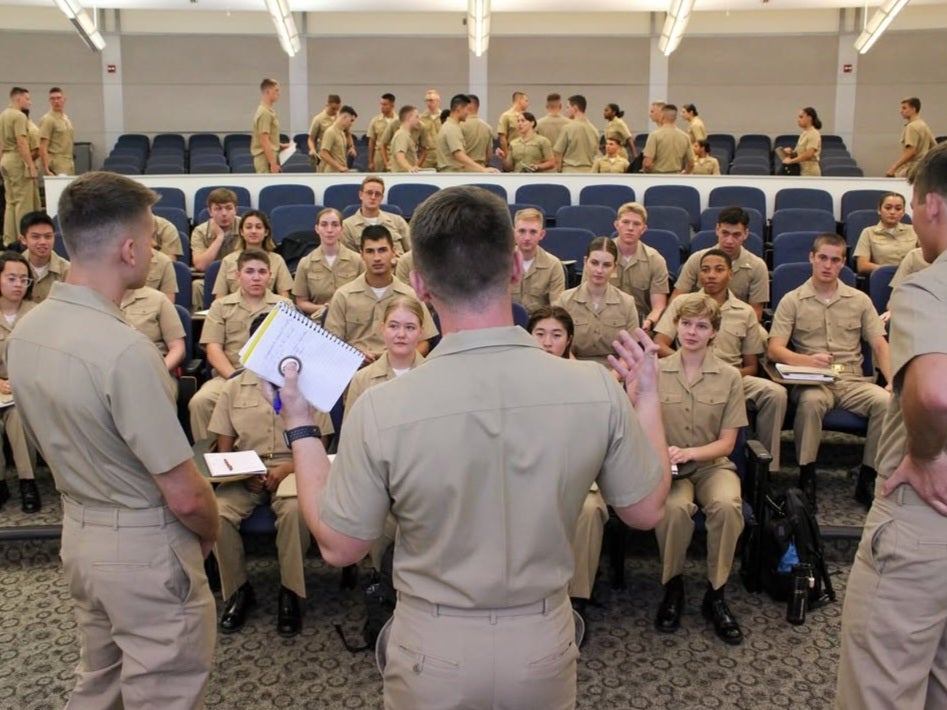 A group of young cadets in beige uniforms sit attentively in a tiered lecture hall. An instructor, also in beige, addresses them, holding a notebook.