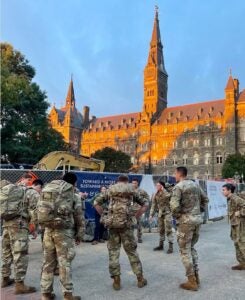 A group of soldiers in camouflage uniforms stand in a circle, talking. Behind them, a historic, sunlit building with tall spires and a clear sky.