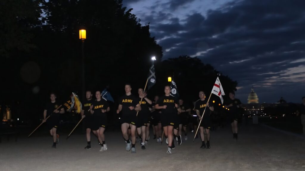 A group of soldiers in black Army T-shirts run along a dimly lit path at dawn, holding flags. The U.S. Capitol is visible in the background under a cloudy sky.