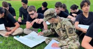 A group of young adults in black shirts sits on grass, studying maps. A person in a U.S. Army uniform is instructing, creating a focused atmosphere.