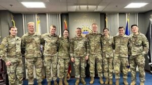 A group of nine soldiers in camo uniforms stand smiling inside a conference room, with a "Joint Staff" sign and flags in the background, conveying camaraderie.