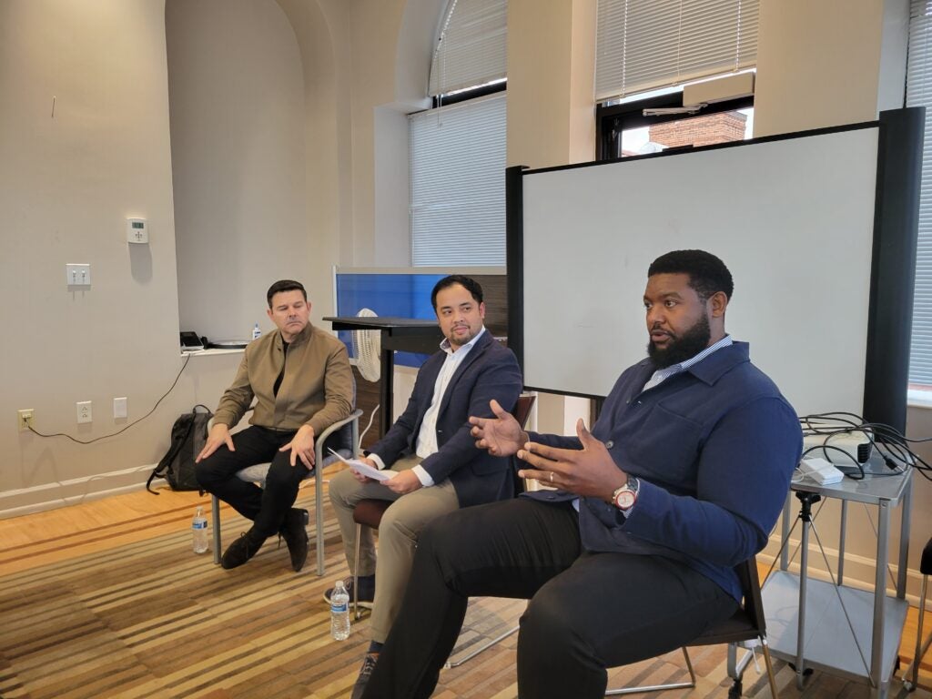 Three men sitting in chairs in front of a screen speaking to an audience