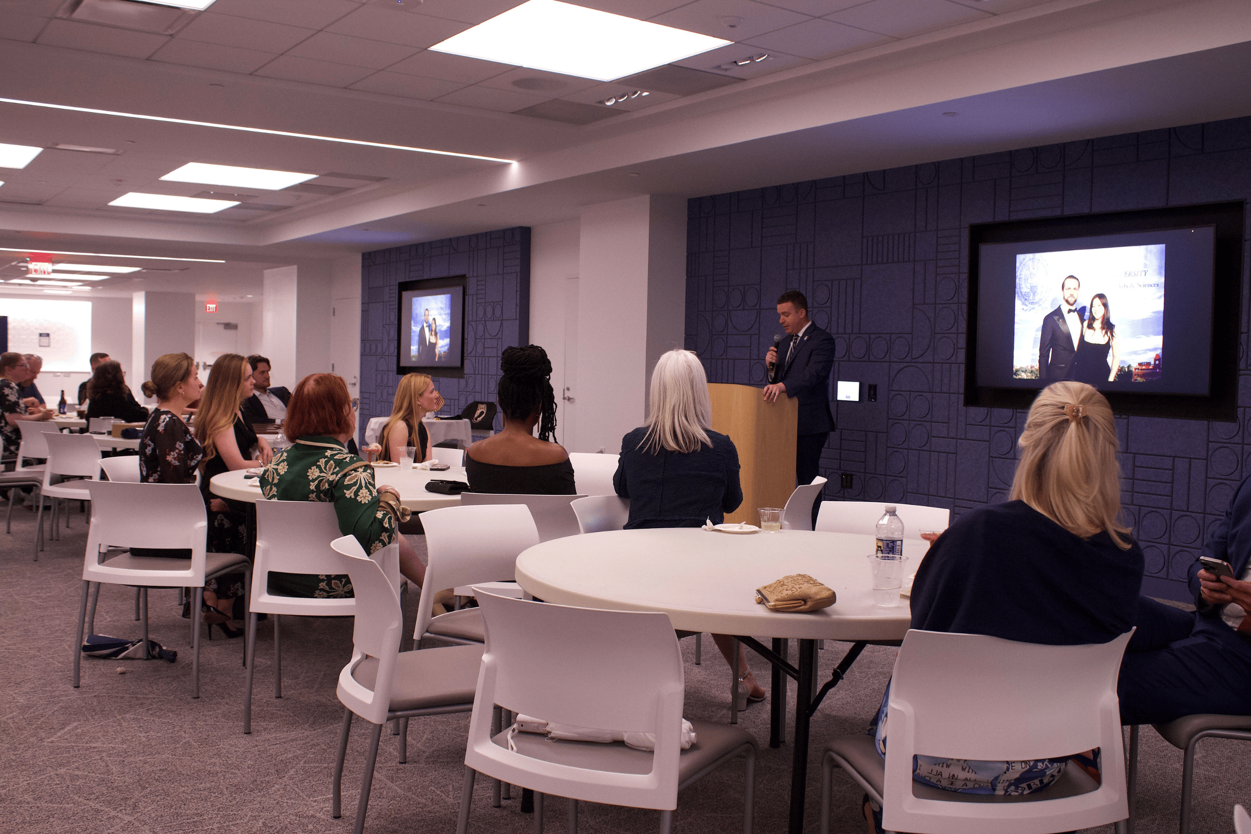 People sitting around white, round tables listen to a man at a podium speak