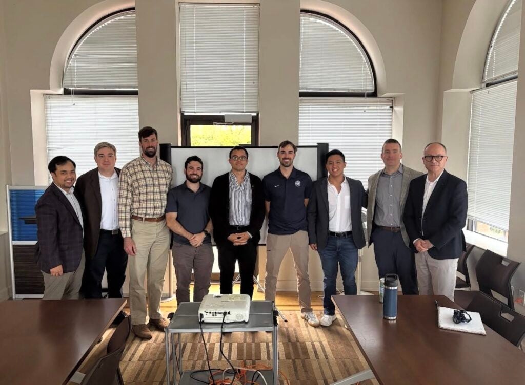 A group of men in suits pose for a picture in front of a room with windows on the wall