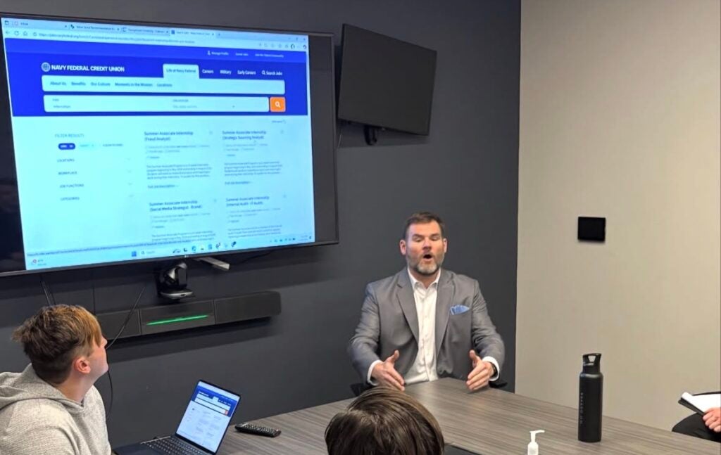 A man presenting to a group sitting around a table in front of a tv screen showing the Navy Federal website