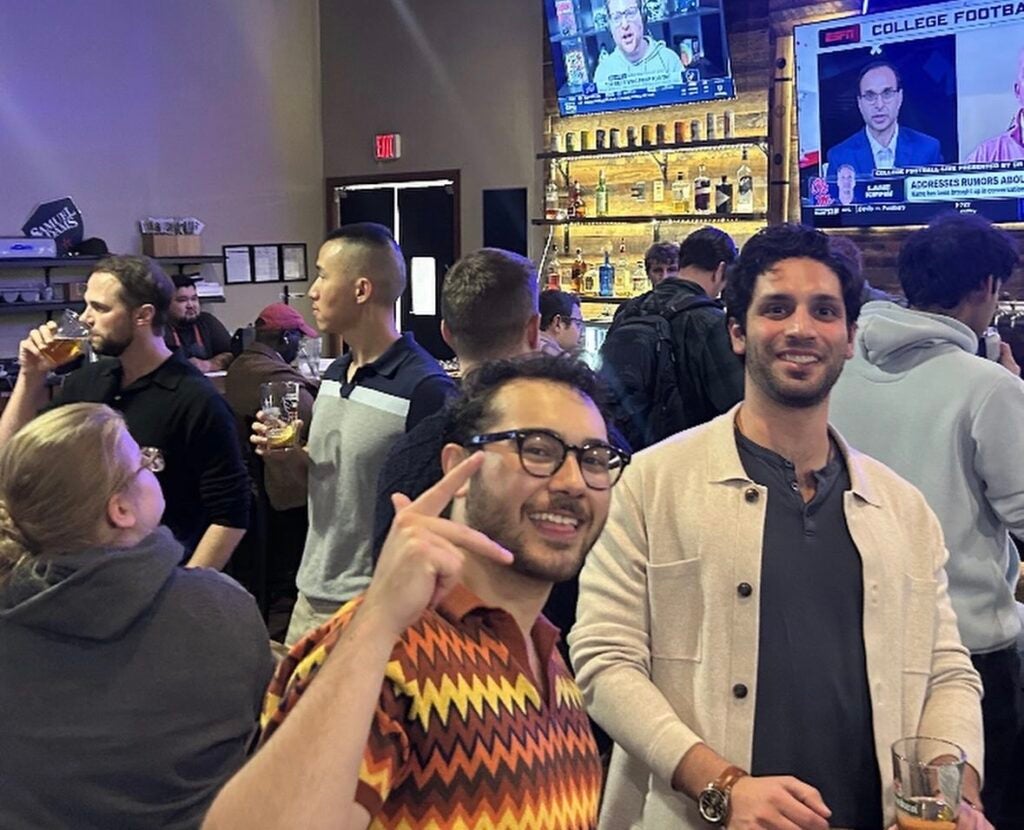 2 men smiling at the camera in front of a group mingling in front of a bar