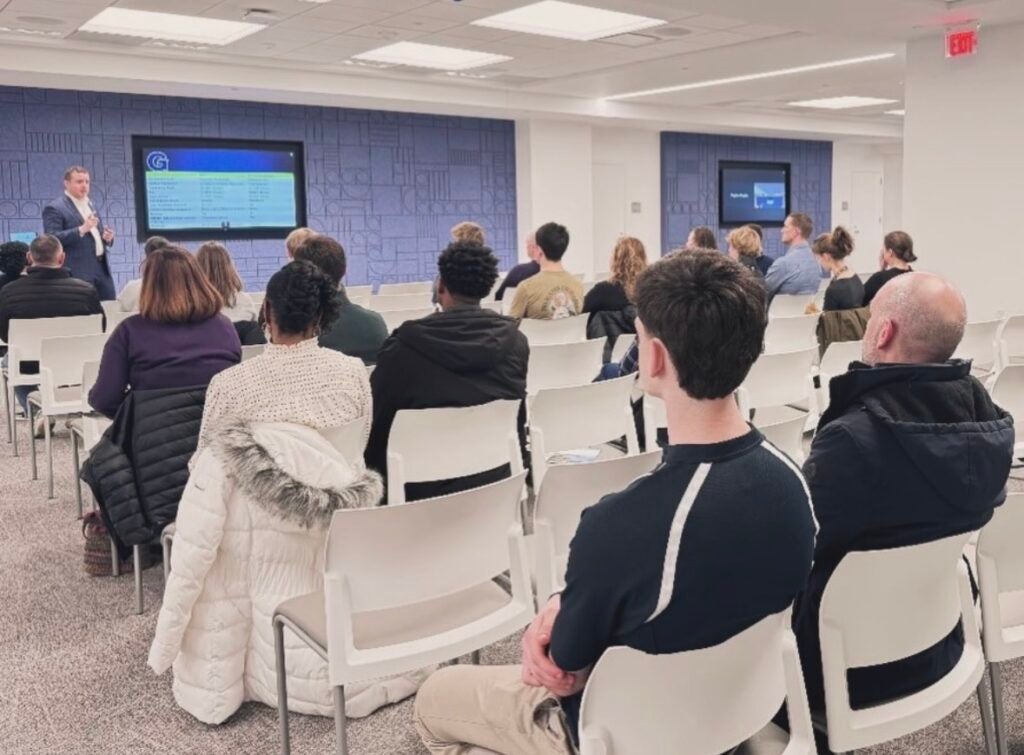 A group of people sit in white chairs listenint to presenters at the front of a room talking in front of TV screens