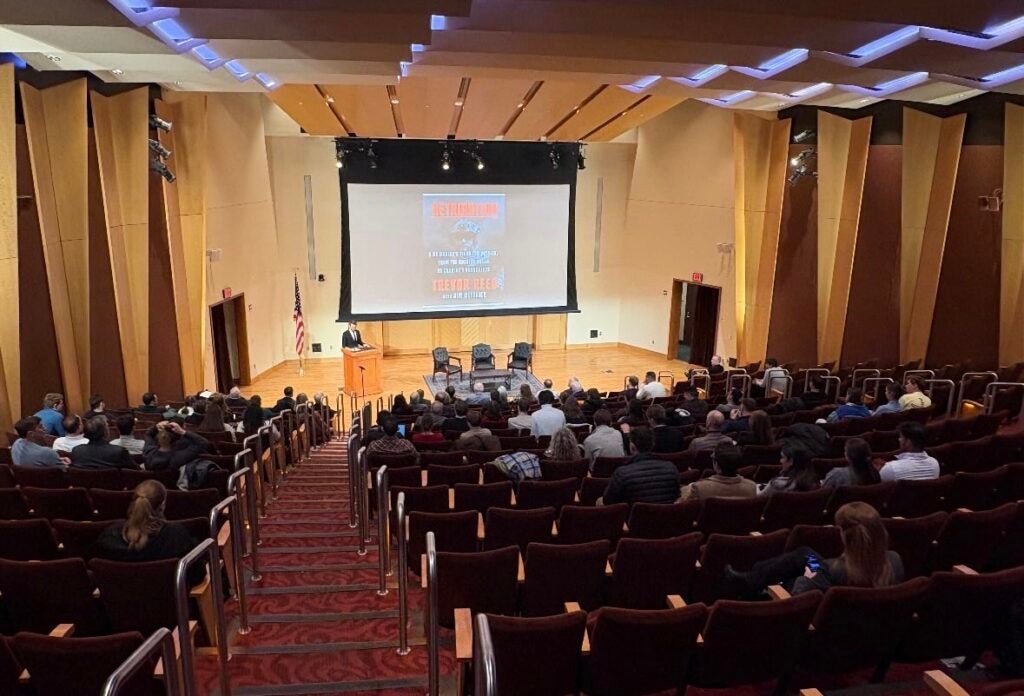 An auditorium full of people looking at a drop down screen and three presenters sitting on a stage