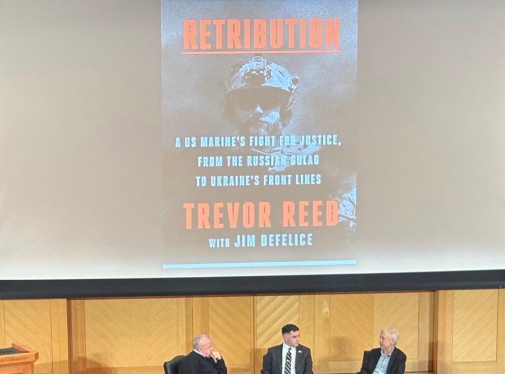 Three men sitting on a stage in a panel in front of a screen with an image of a book cover titled 'Retribution'