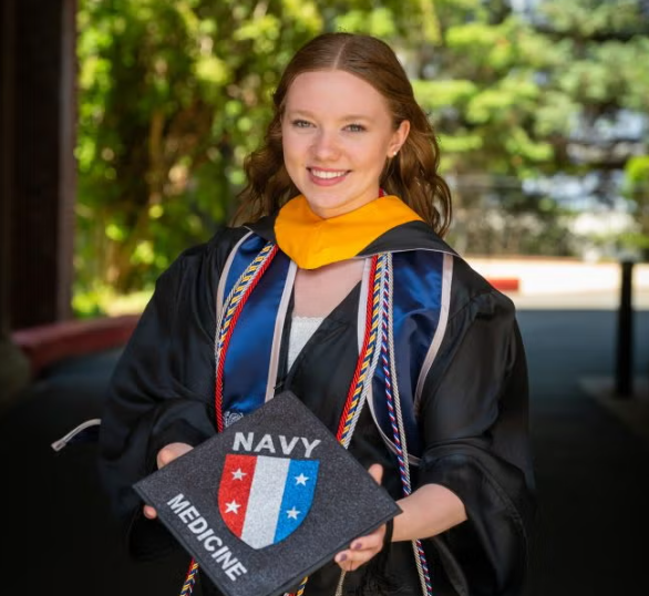 A blond woman in graduation regalia holding a cap that says 'navy medicine'