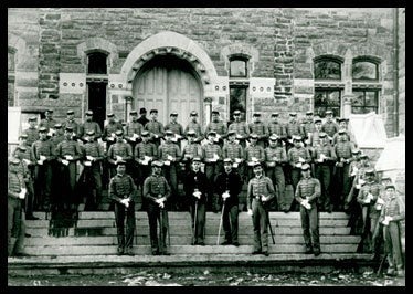 Cadet soldiers in uniform standing in front of a brick building in black and white