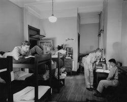 Students in uniform in a dorm room with bunk beds and desks studying